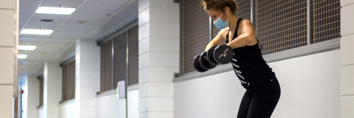 Female lifting dumbells in a black outfit and a disposable face covering.