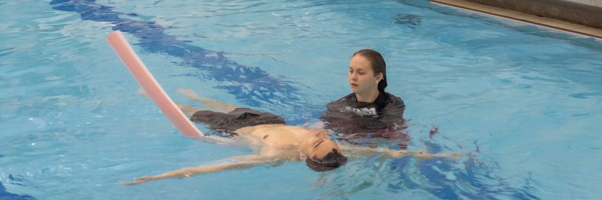 Swim lesson participant floats on their back supported by a pool noodle and instructor. 