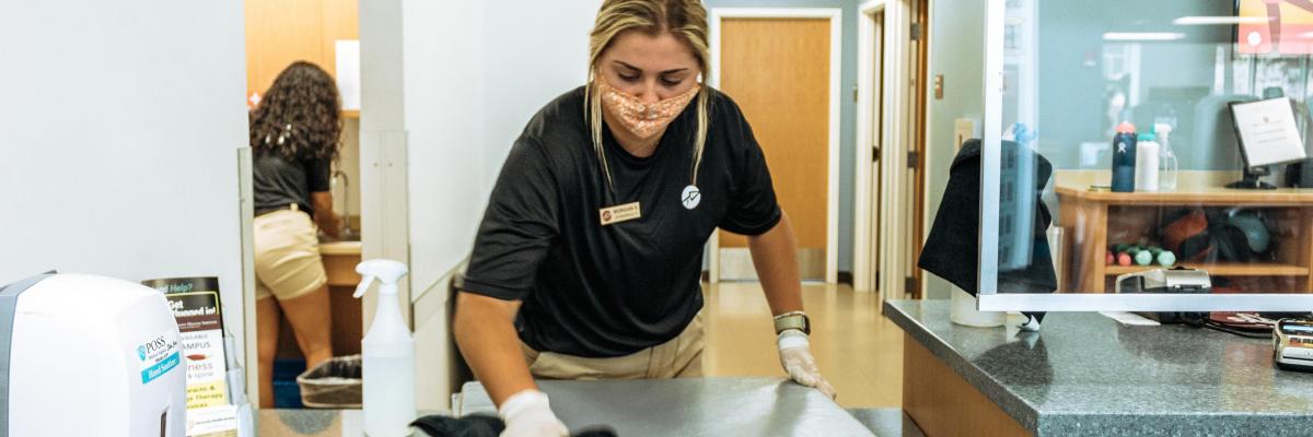A woman in a mask and gloves wipes down a fitness mat at the Leach center Fitness desk