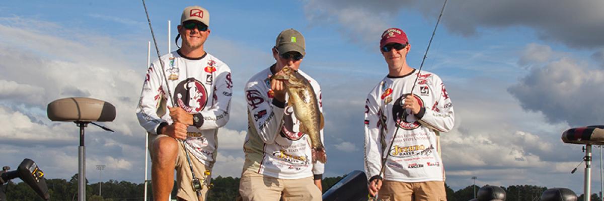 Three males on a boat. First male holding fishing pole and smiling, second male kissing caught fish, third male holding fishing pole and smiling. 
