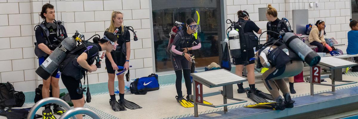multiple students with oxygen tanks on backs learning from scuba instructor