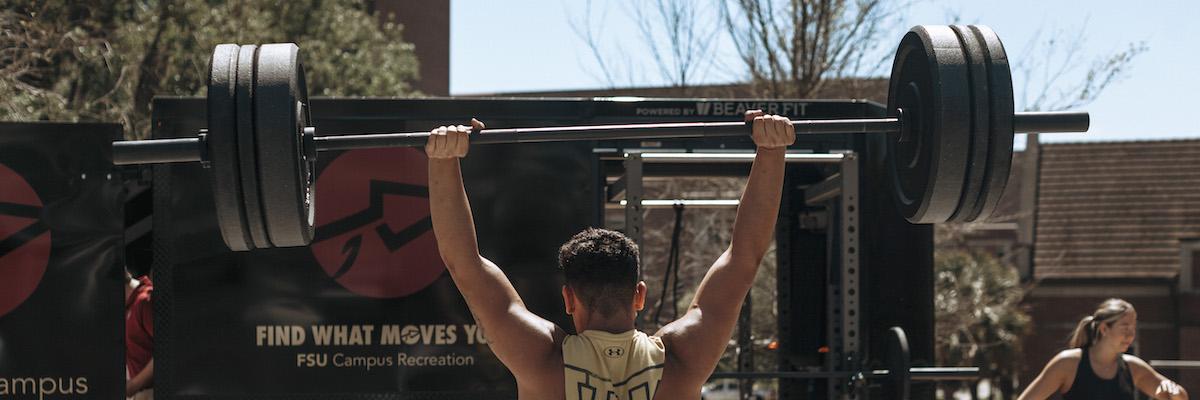 Male student lifting a barbell with weights above his head, facing away from the camera