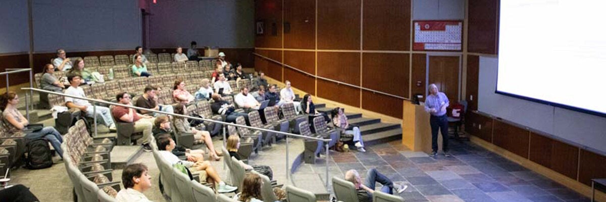 Presenter speaking to an audience in a tiered lecture hall during a seminar or workshop.