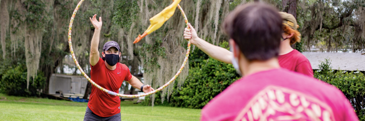 Masked participate throws a rubber chicken through a hula hoop. 