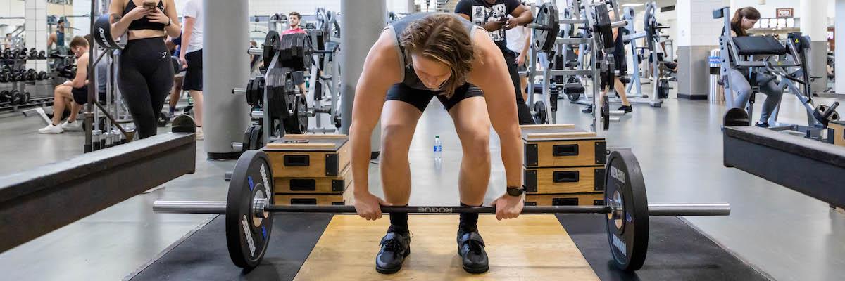 Male student on lifting platform bending down to pick up barbell