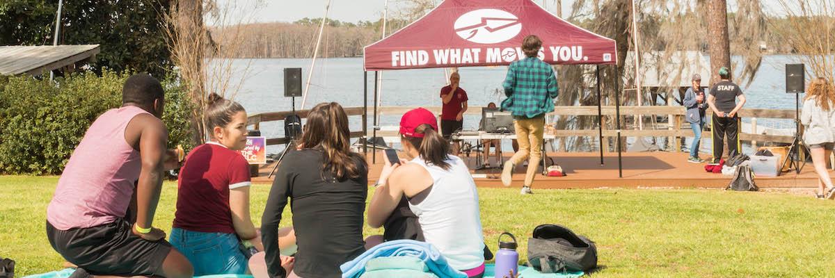 Four students sit on a blanket in the grass looking out at the lake, with a Find What Moves You tent in their view