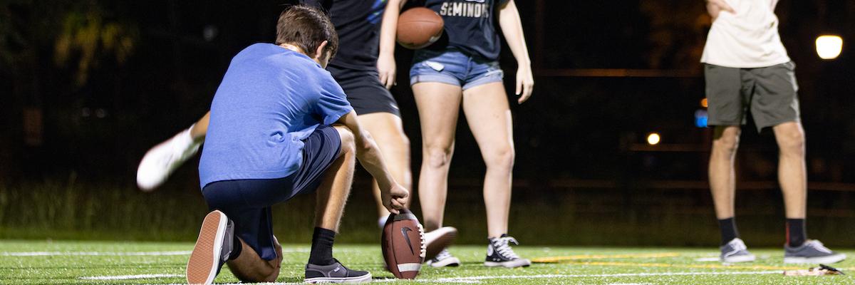 male student kneeling and holding a football for another student to kick it