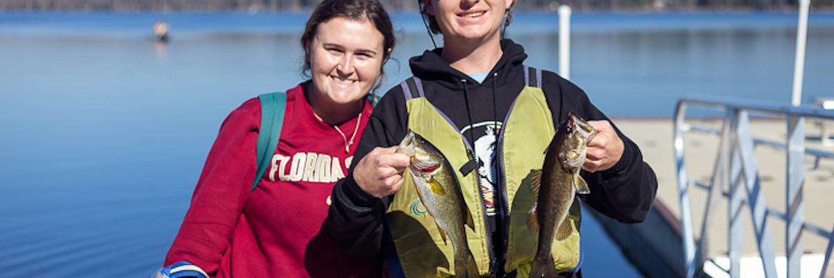 male and female student holding up fish in front of lake