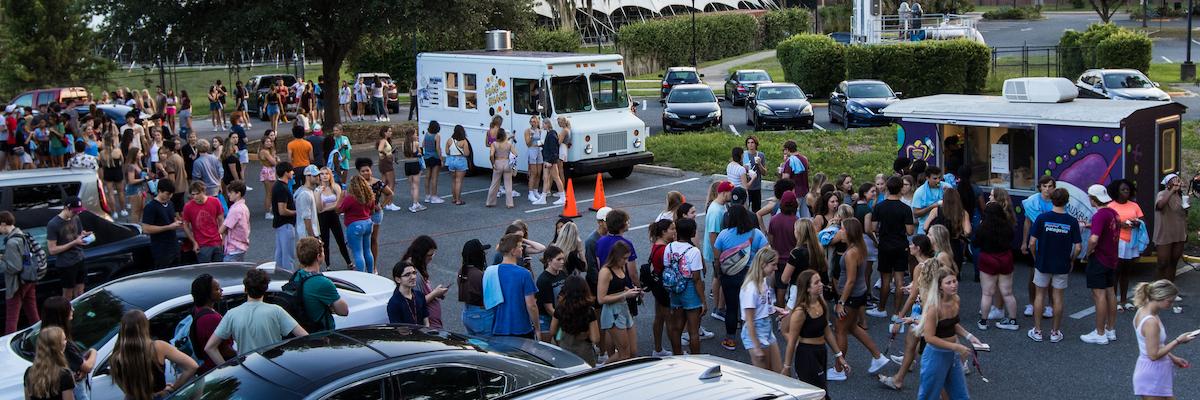 Many students surrounding two food trucks in a parking lot